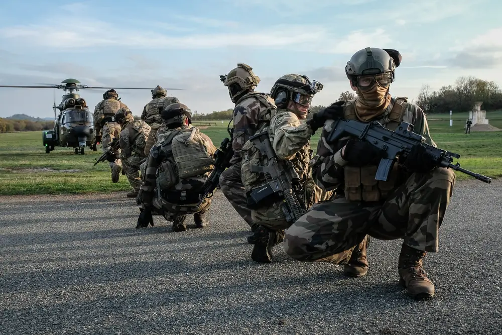 Un groupe de soldats en tenue de camouflage et équipement de combat est accroupi en ligne sur une route, avec des armes. Un hélicoptère militaire est visible en arrière-plan sur un terrain herbeux sous un ciel nuageux.