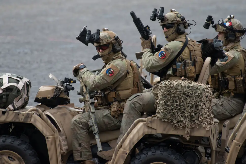 Trois soldats équipés de tenues et de casques militaires, avec des dispositifs de vision nocturne et des armes, sont assis sur des véhicules tout-terrain de couleur sable. Le soldat au premier plan tient son arme prête. Ils semblent être en mission ou en exercice.