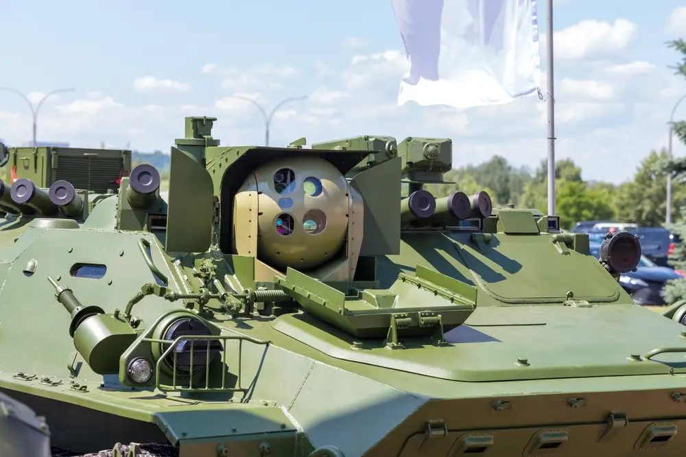 Un gros plan d'un véhicule militaire blindé de couleur verte, équipé d'un système optronique sphérique monté à l'avant, avec plusieurs lentilles visibles. En arrière-plan, un ciel bleu et des éléments d'un événement en extérieur.