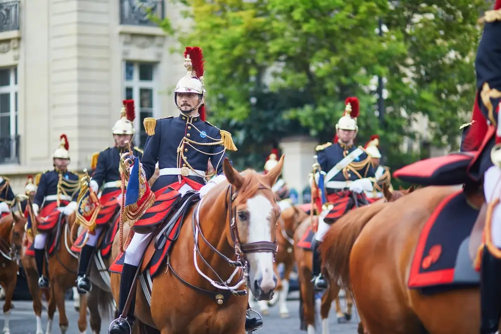 Un groupe de cavaliers de la Garde républicaine à cheval, vêtus de leur uniforme de cérémonie, incluant un casque avec une crinière rouge, une veste bleue ornée de galons dorés et un pantalon blanc, défilant dans une rue.