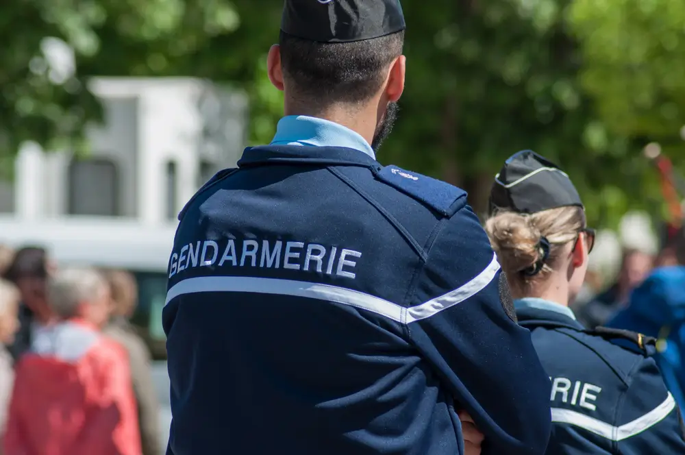Deux gendarmes, un homme et une femme, vus de dos. L'homme, au premier plan, porte une veste de gendarmerie bleu marine avec l'inscription 'GENDARMERIE' en lettres blanches sur l'épaule, illustrant l'uniforme typique de la gendarmerie nationale en France.