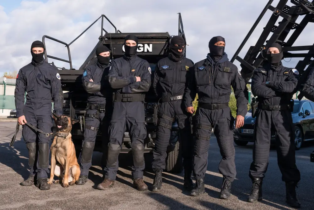 Plusieurs gendarmes du GIGN, vêtus d'uniformes d'intervention noirs, cagoulés et équipés de masques de ski ou de lunettes, posent devant un véhicule de transport blindé noir. Un chien de travail est également présent.