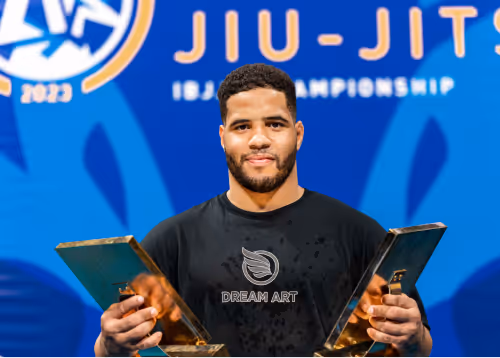 A Jiu-Jitsu champion holding two trophies on stage in front of a championship banner