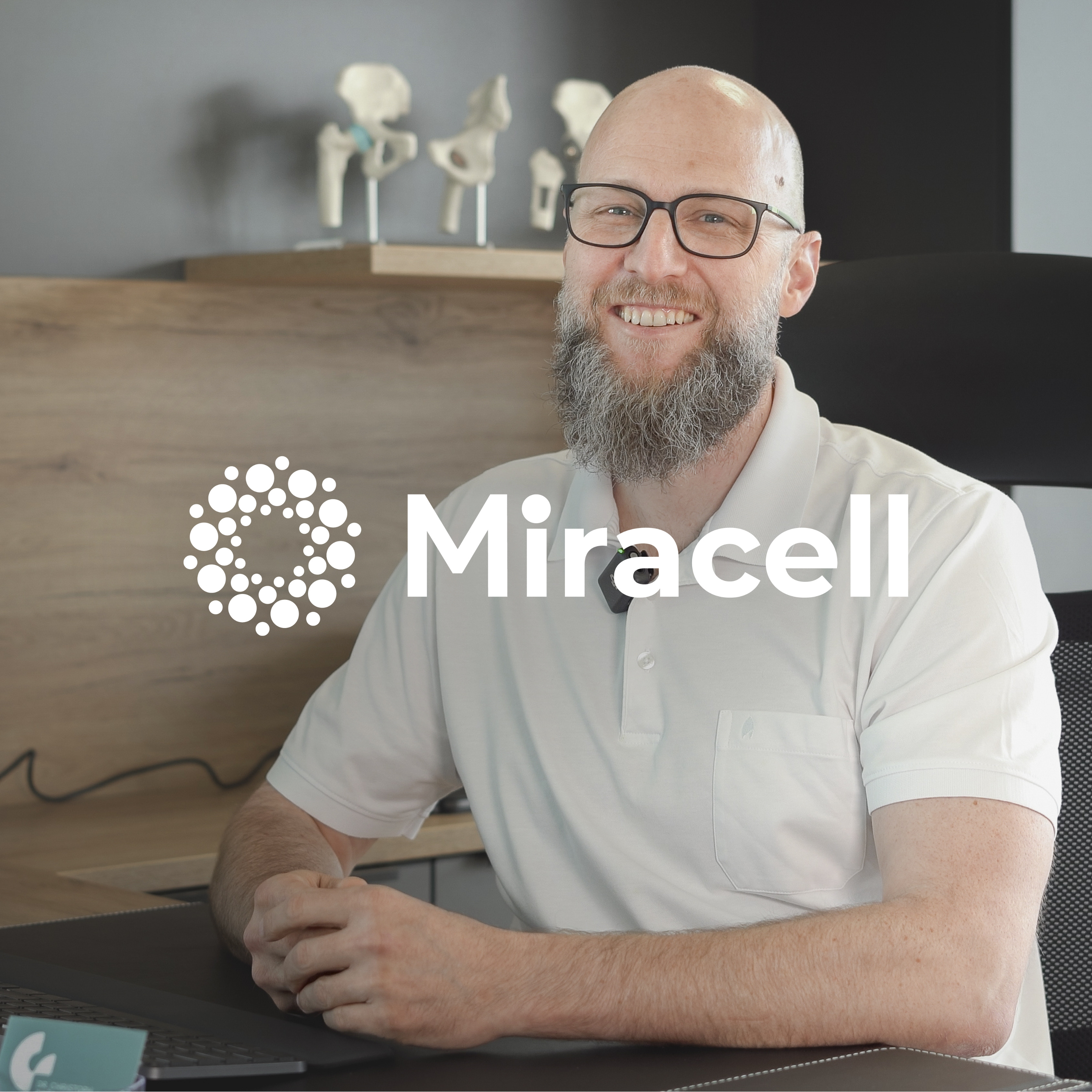 lächelnden Mann mit Brille und Vollbart. er sitzt in einer Ordination an einem Schreibtisch. über dem Bild liegt das Miracell Logo. Dr. Gröbner