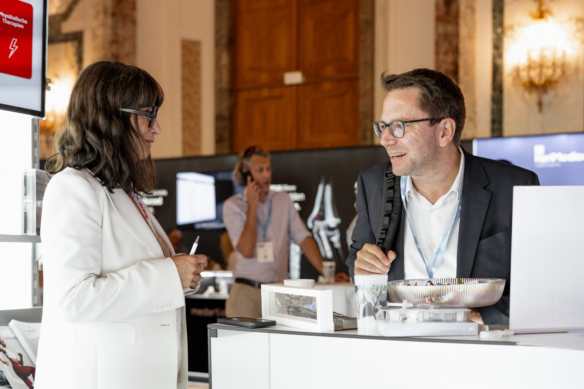 Eine Frau im weißen Blazer spricht mit einem lächelnden Besucher am Messestand.
