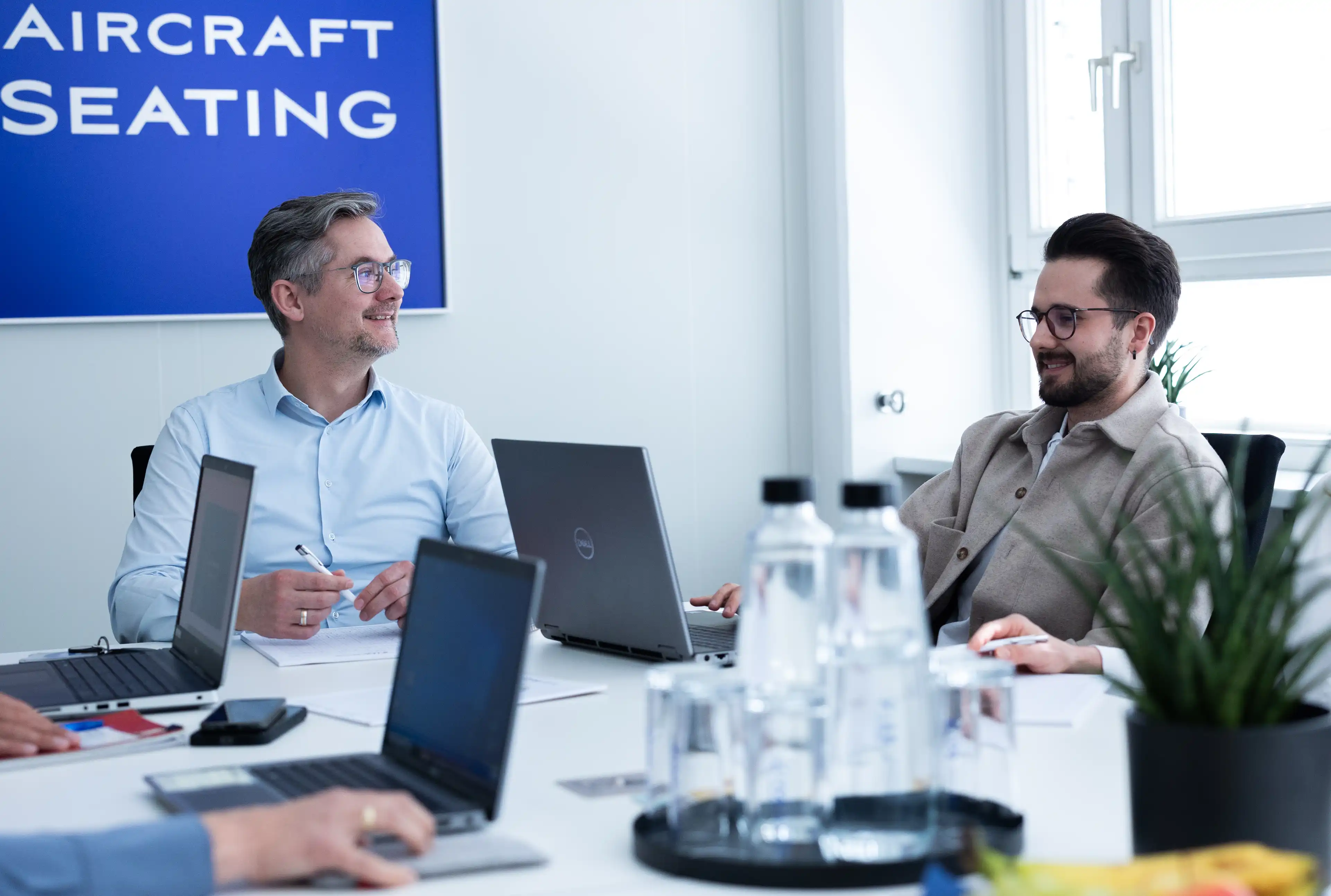 Zwei Männer mit Brille unterhalten sich bei einem Meeting mit geöffnetem Laptop und Wasserflaschen auf dem Tisch. Es ist am Rand erkennbar, dass noch mehr Personen am Tisch sitzen, da Arme erkennbar sind.