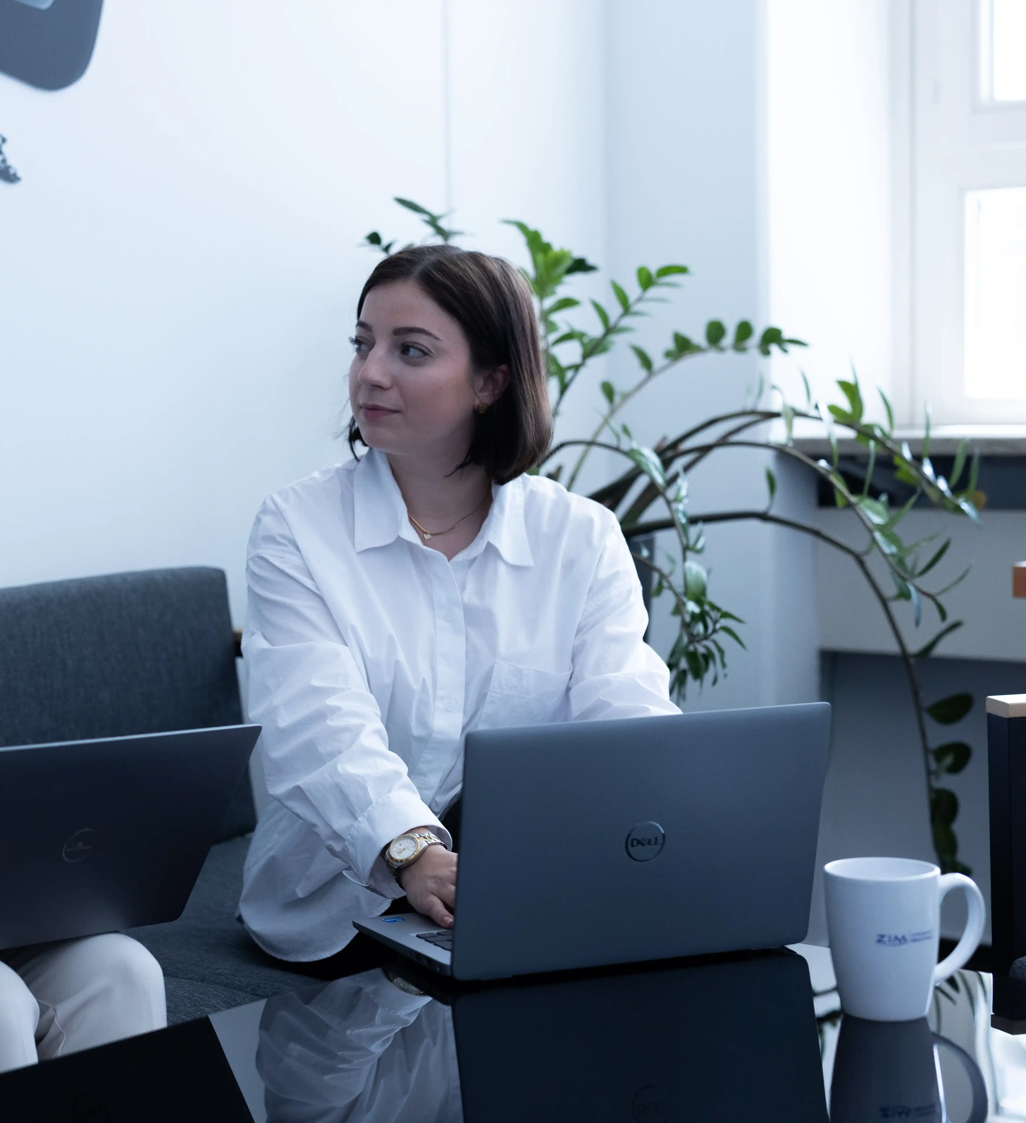 Frau mit weißer Bluse arbeitet an einem Laptop in einem modernen Büro mit Pflanze im Hintergrund.