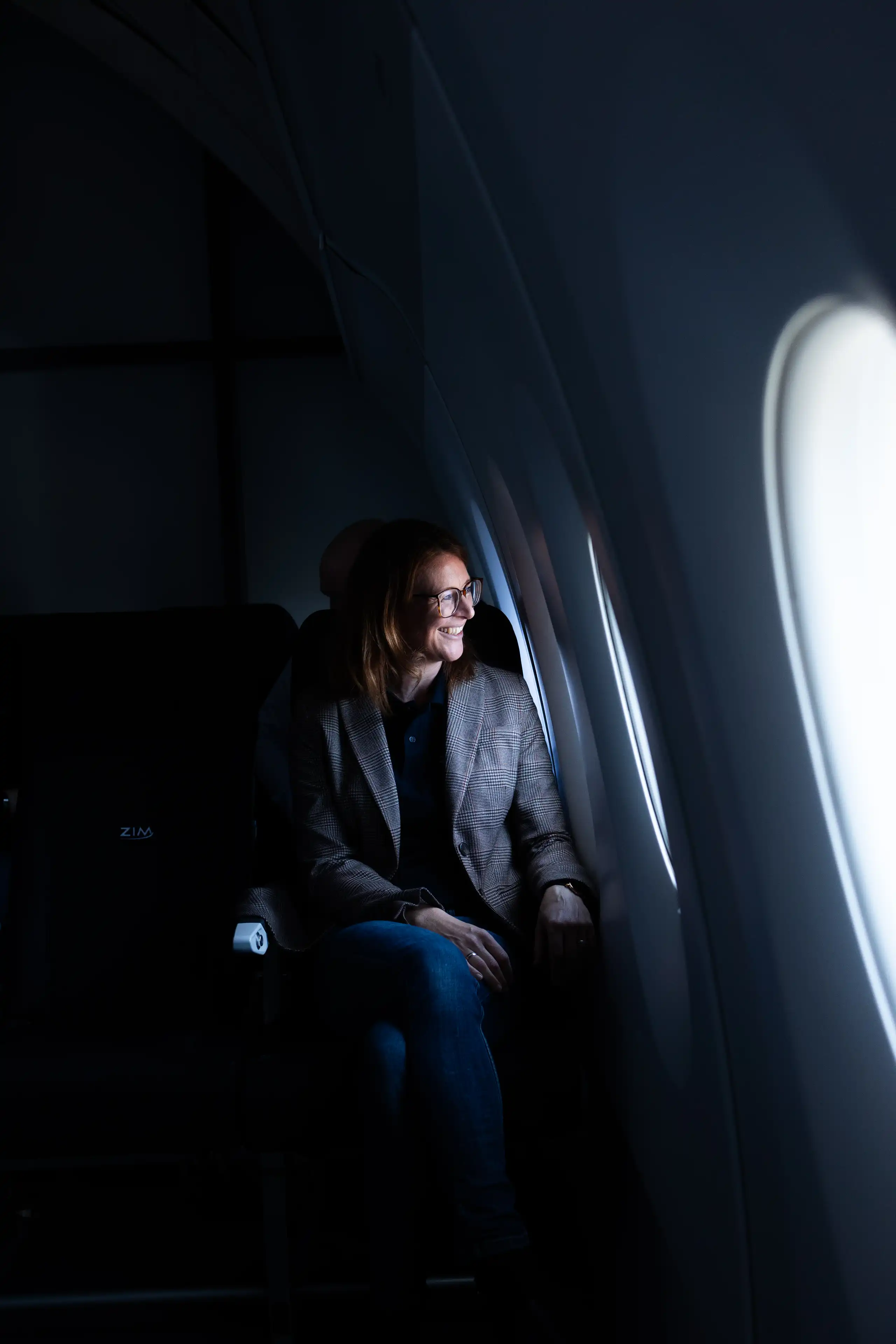 Frau mit Brille sitzt lächelnd am Fenster eines Flugzeugs und schaut hinaus.