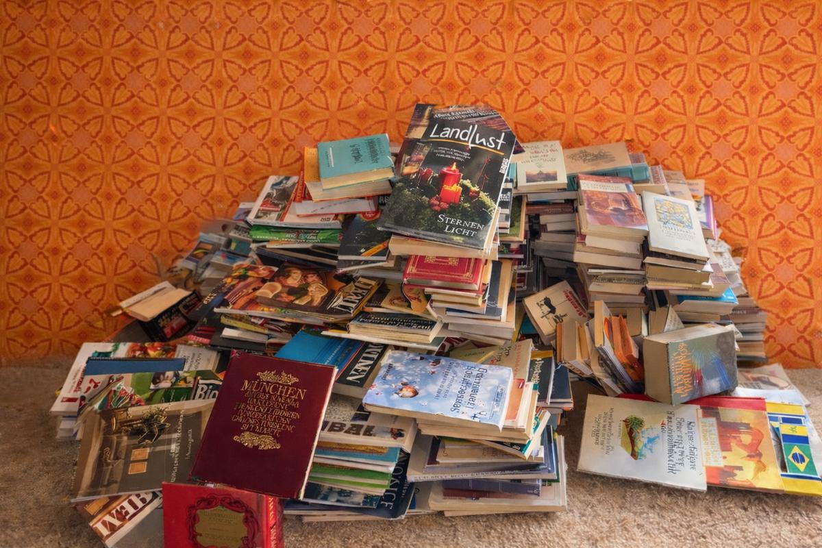 Large pile of assorted books stacked on a beige carpet against an orange patterned wall.