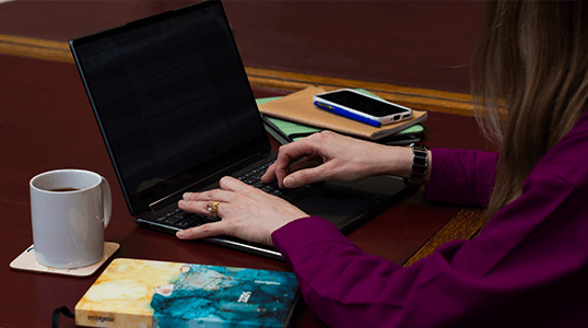 Person typing on a laptop with a coffee mug and notebooks nearby on a wooden table.