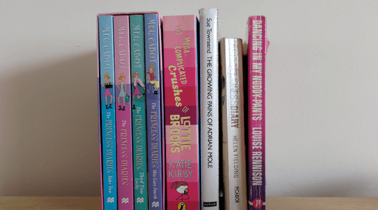A row of eight colorful books standing upright on a wooden surface against a plain wall.