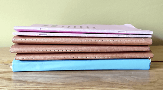Stack of four closed notebooks with pink, two brown, and blue covers on a wooden surface against a pale green background.
