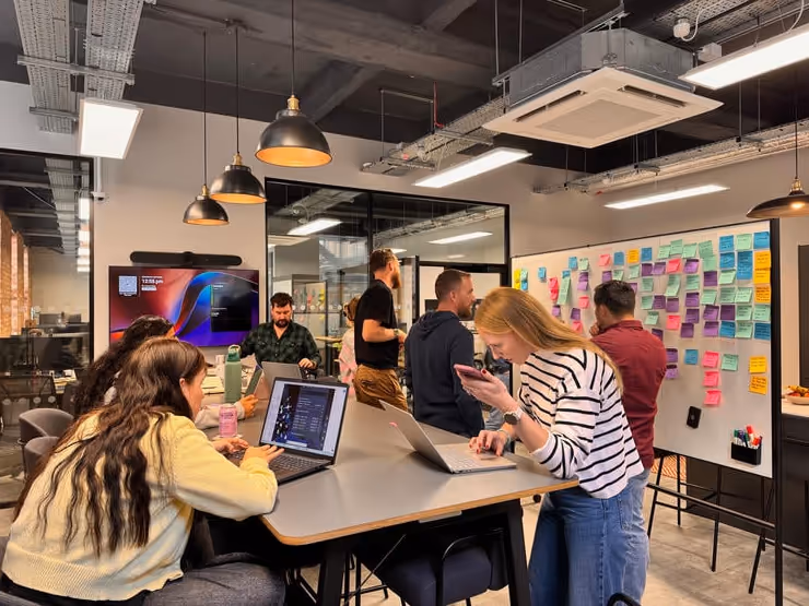 Group of coworkers collaborating in a modern office with laptops and a whiteboard covered in colorful sticky notes.