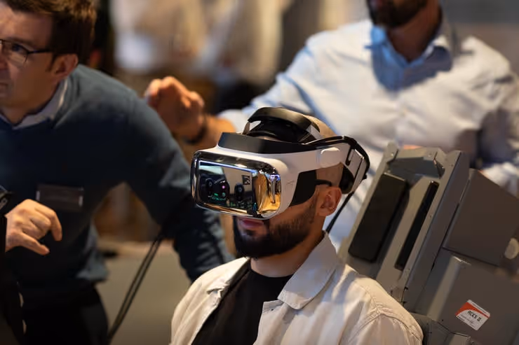 Man wearing a VR headset sitting while two other men observe and interact nearby.