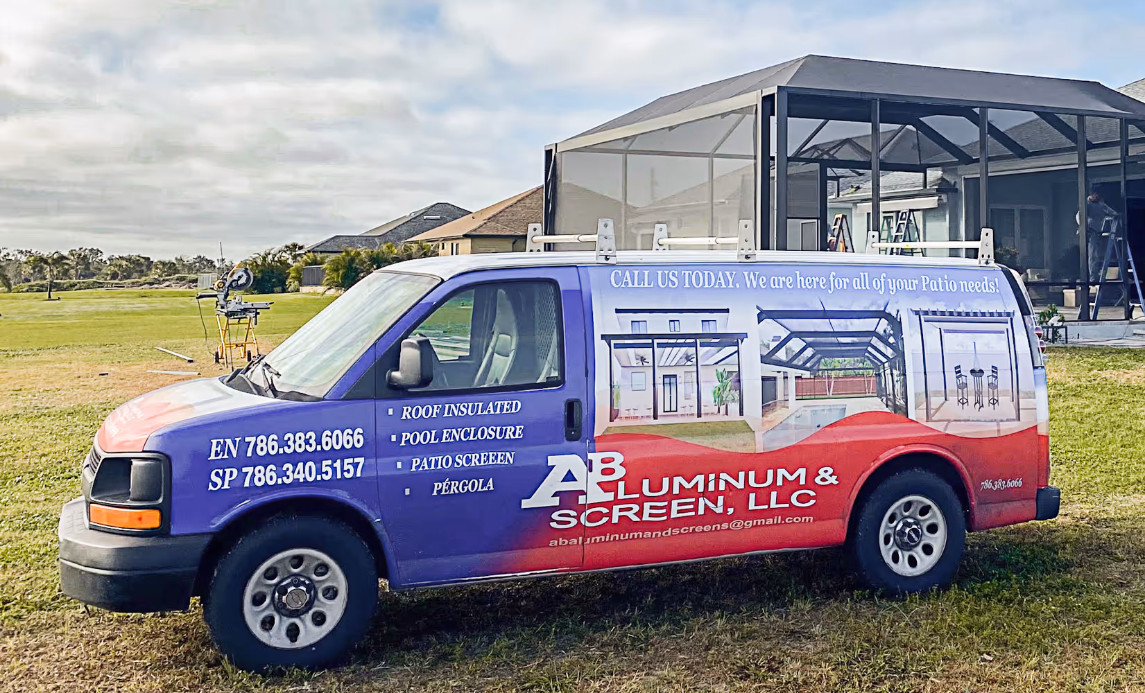 Side view of a blue and red aluminum and screen service van parked on grass with patio enclosures in the background.