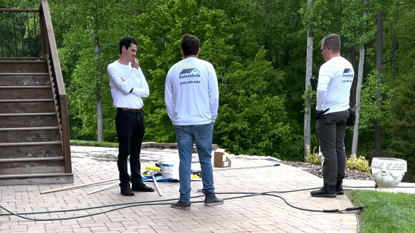 Three men in white Estatehub shirts standing on a stone patio with tools and greenery around.