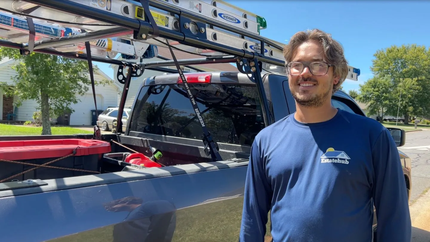 Man with glasses wearing blue Estatehub shirt stands smiling beside a truck with ladders on a sunny street.