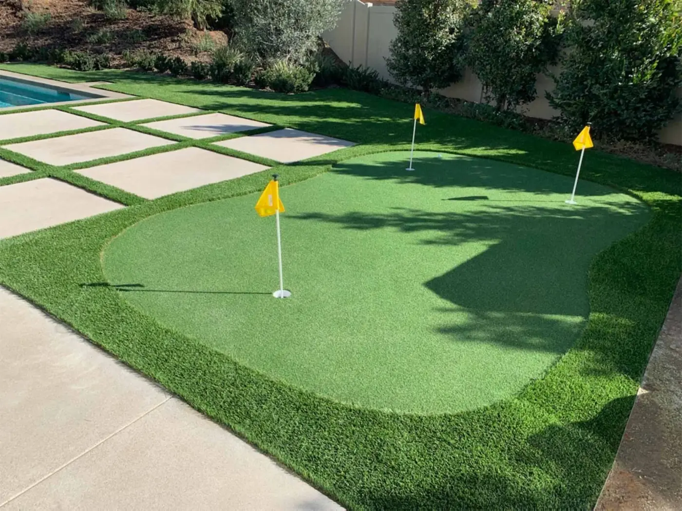 A backyard putting green with yellow flags and paver steps beside a pool, installed by United Turf
