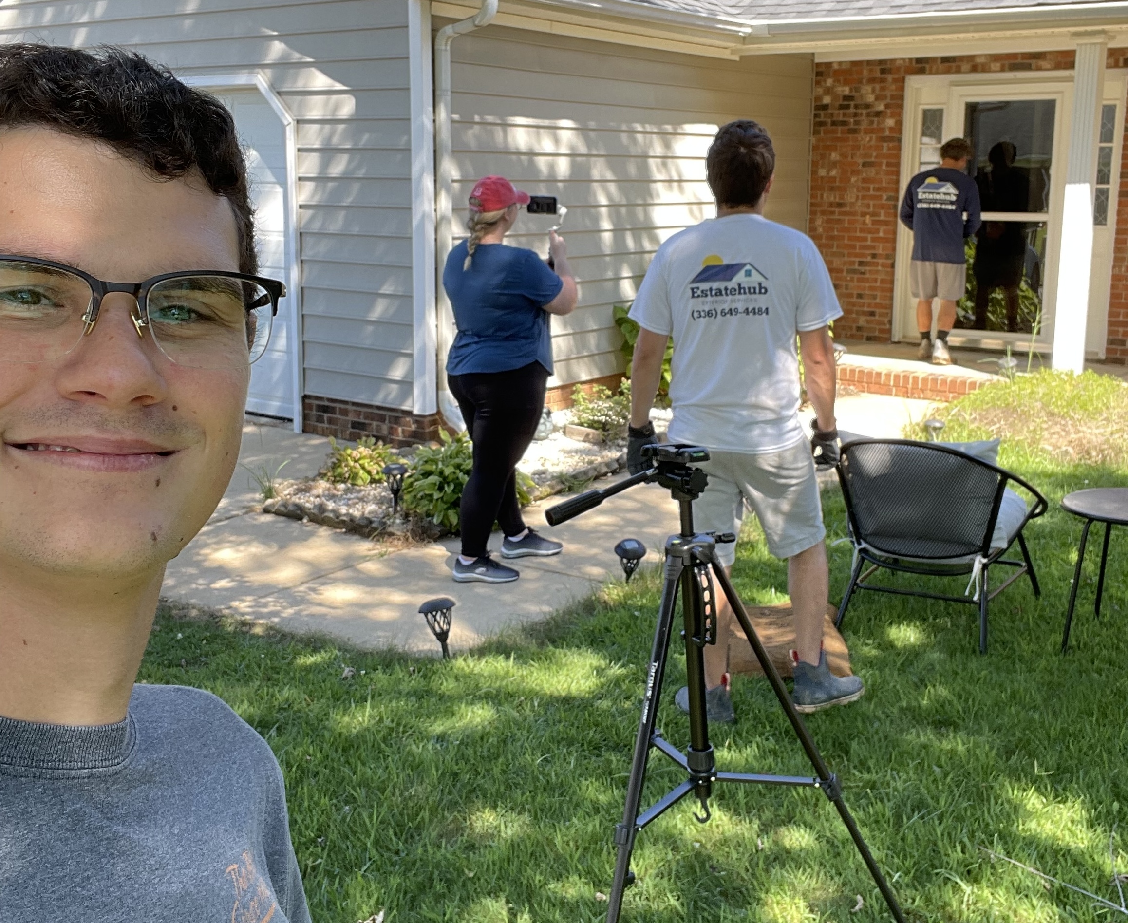 Team members filming content in a home's front yard, with a phone on a tripod and a smiling man in glasses in the foreground.