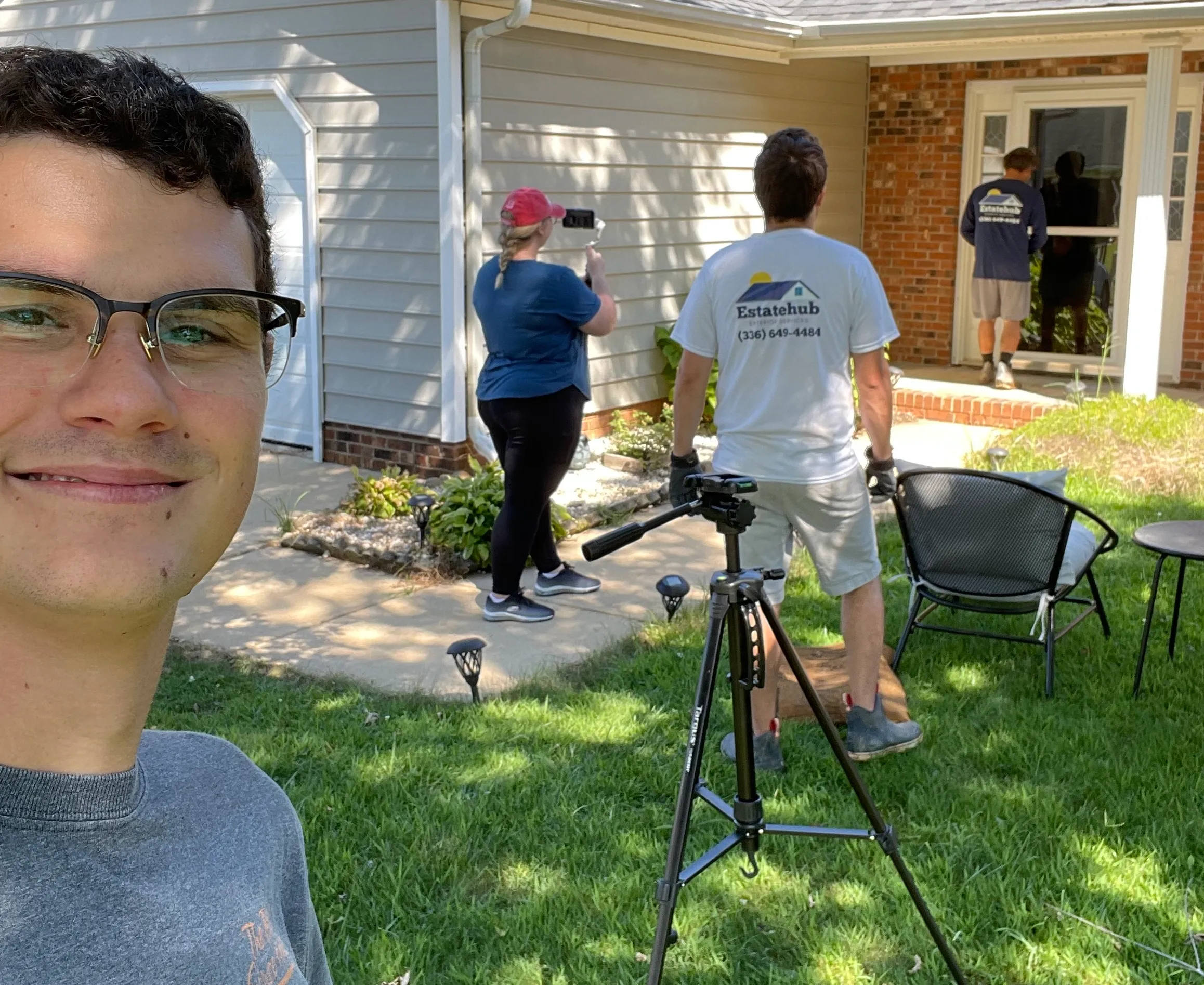 Team members filming content in a home's front yard, with a phone on a tripod and a smiling man in glasses in the foreground.