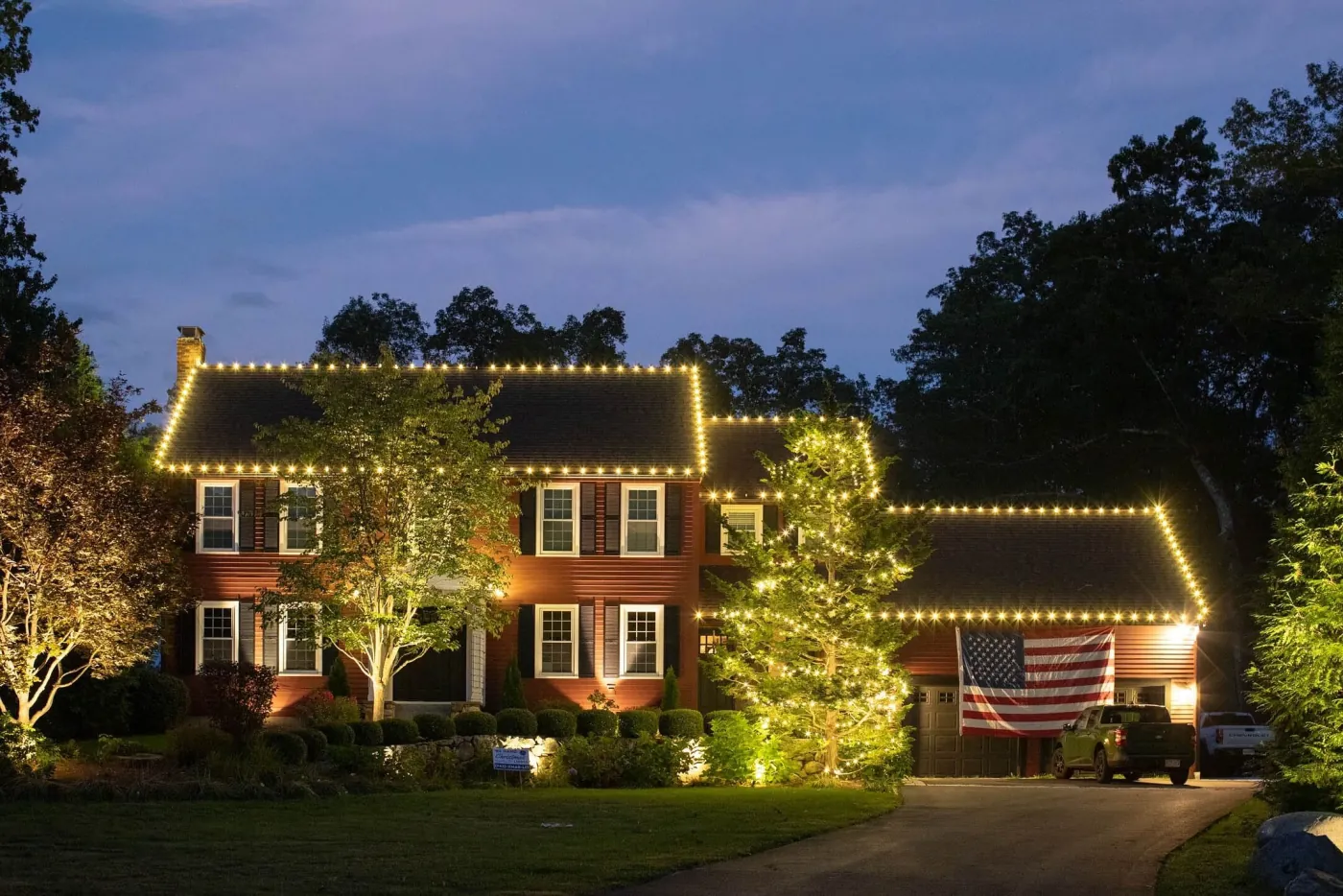 A brick two-story home at dusk with white lights along the roofline and a lit tree, lit by ViewCrew