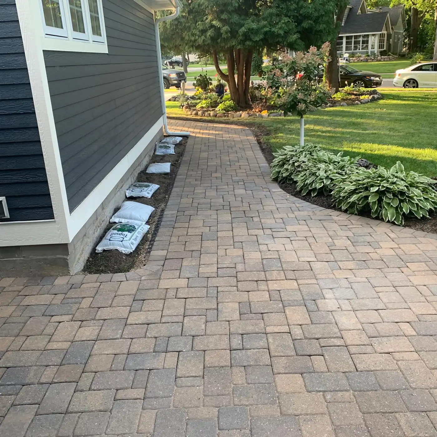 A curved paver walkway beside a house and hosta garden, installed by Purpose Landscaping