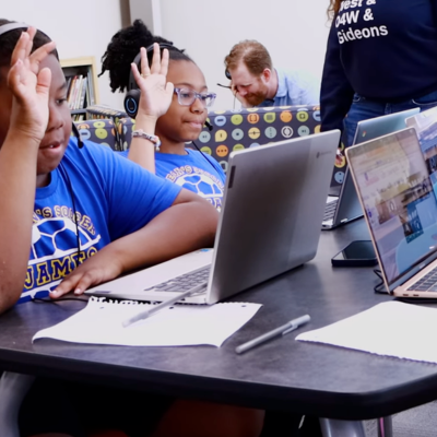 Two students one raising their hand, sitting at a table using laptops with notebooks and pens in front of them