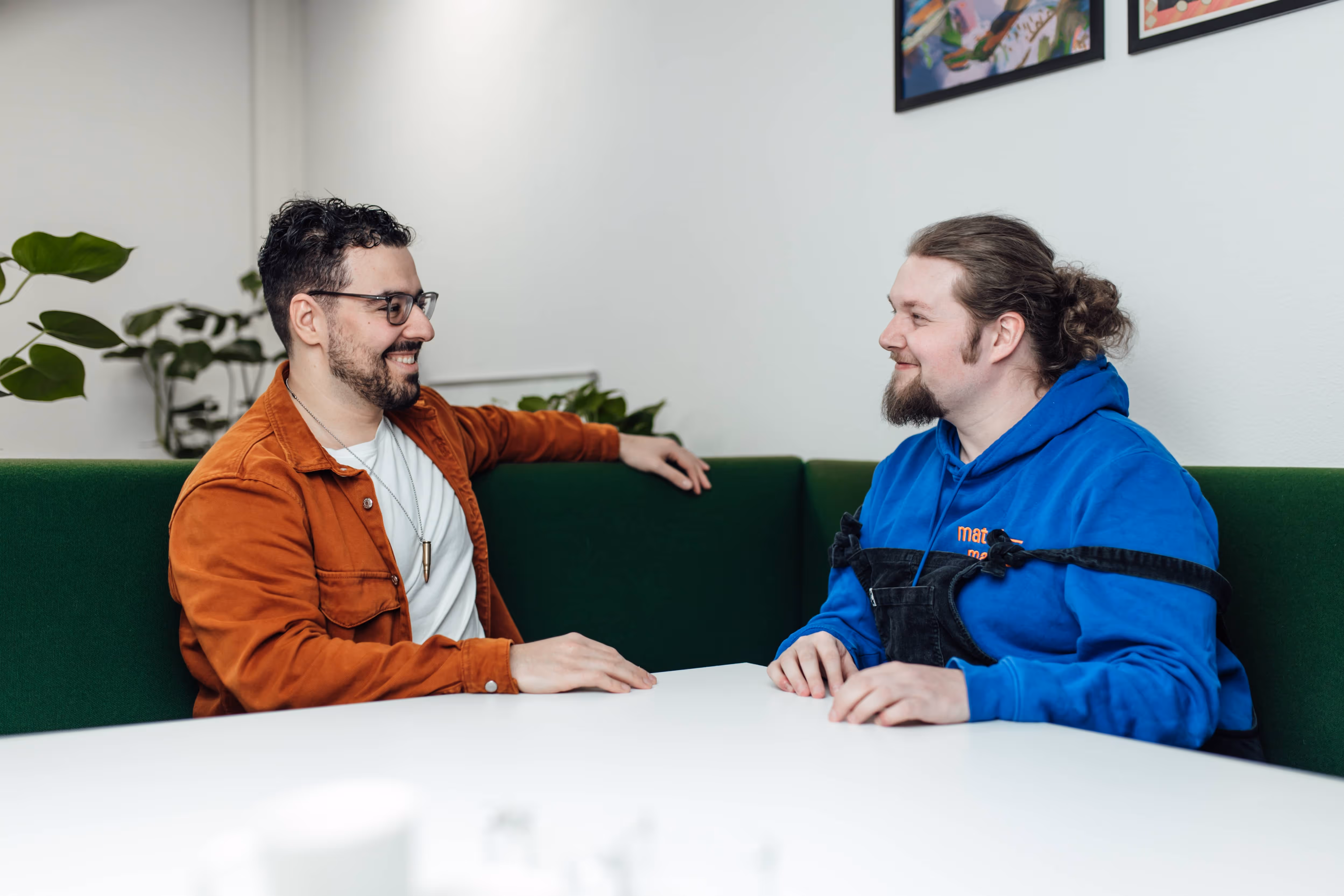 Twee mannen glimlachend zittend aan een witte tafel met groene bank in een lichte kamer.