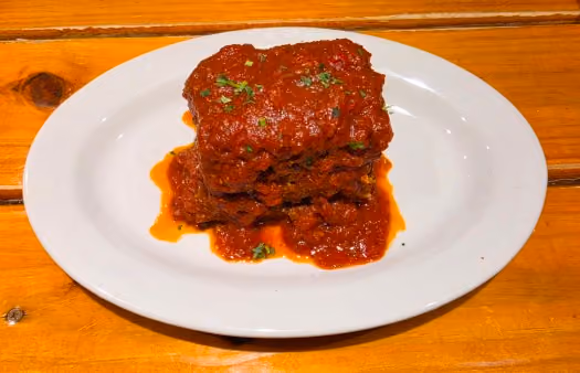 Three stacked pieces of lasagna covered in tomato sauce and garnished with herbs on a white plate, placed on a wooden surface.