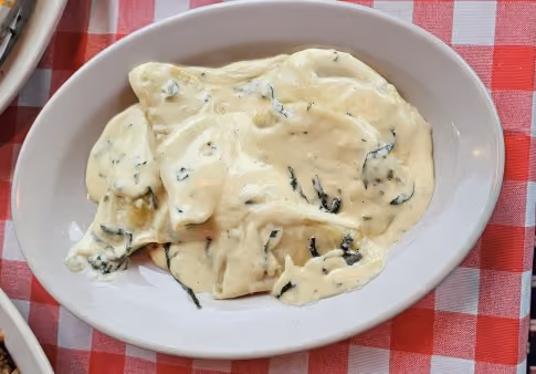White oval dish with three ravioli pieces covered in creamy white sauce with herbs on a red and white checkered tablecloth.
