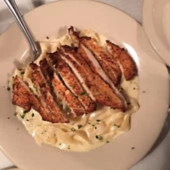 Sliced grilled chicken breast served over creamy fettuccine Alfredo pasta on a round light-colored plate with a fork.