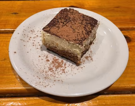 Square piece of tiramisu dessert dusted with cocoa powder on a white plate on a wooden table.