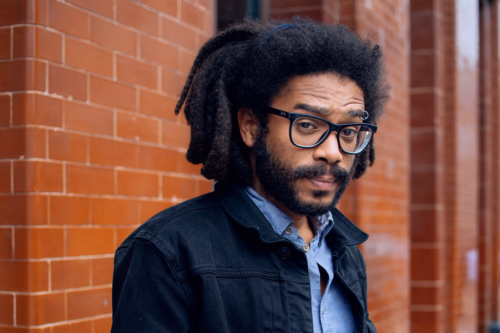 Confident man with dreadlocks and glasses standing by a brick wall, wearing a denim shirt and black jacket