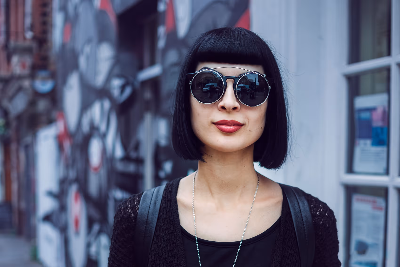 Stylish woman with black bob haircut, round sunglasses, and red lipstick, standing in front of street art