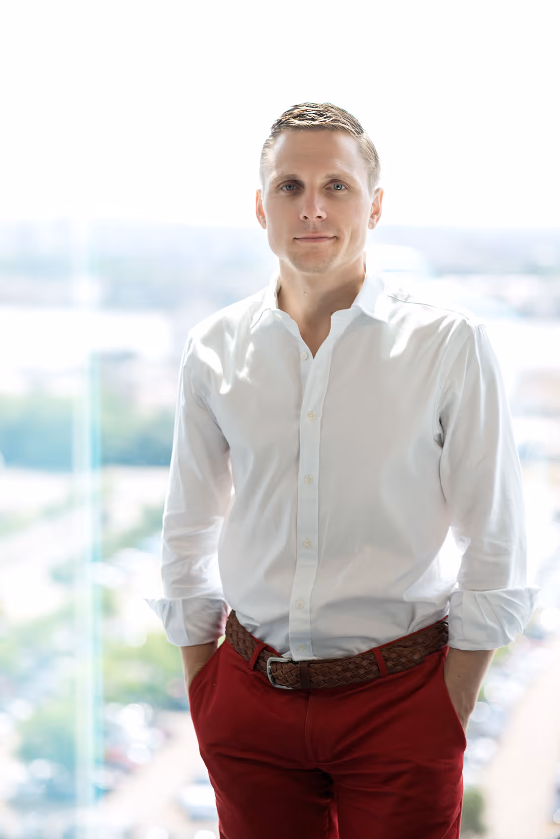 Man in white shirt and red trousers standing by a window with a bright city view in the background