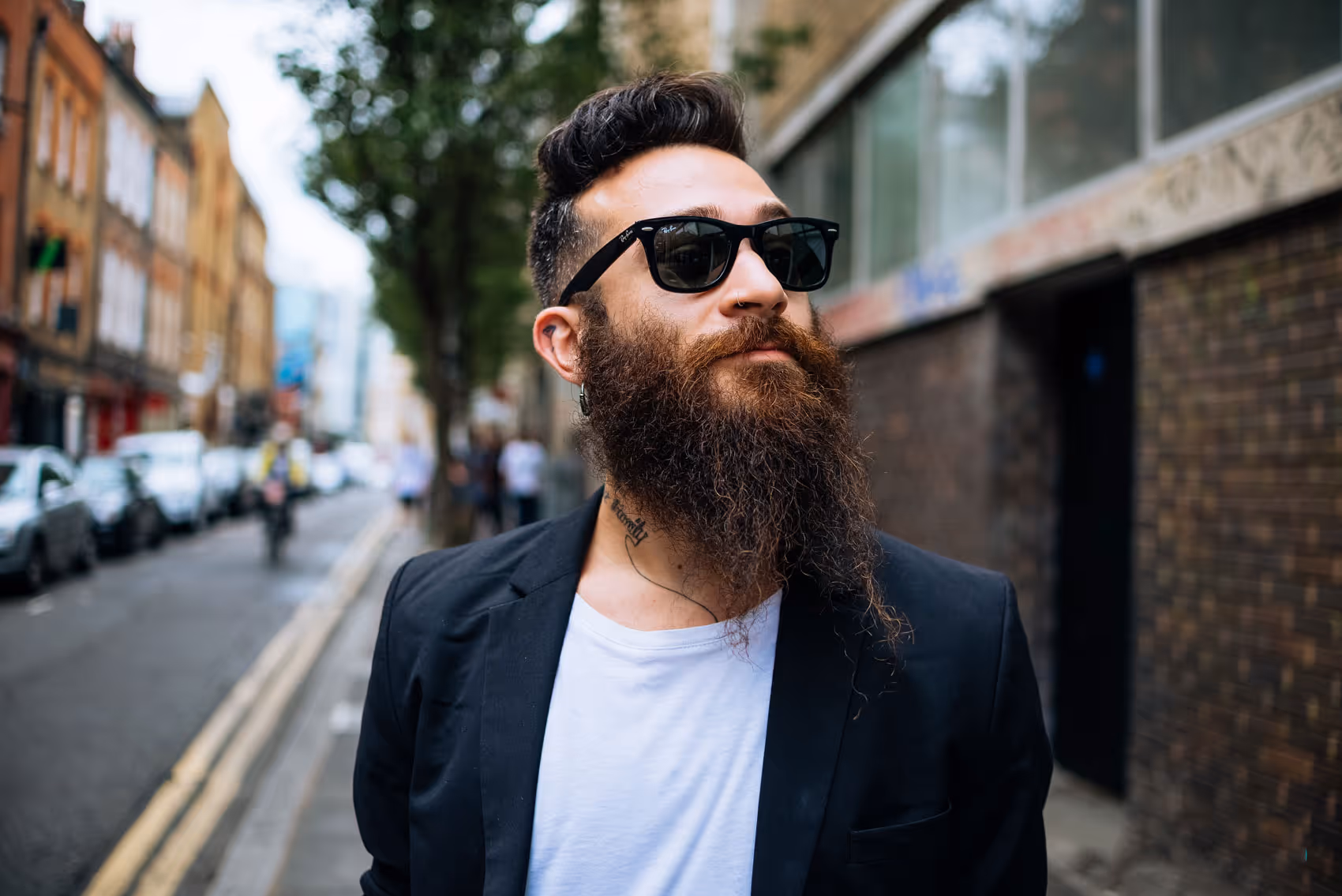 Bearded man with sunglasses and tattoos wearing a blazer, standing on an urban street