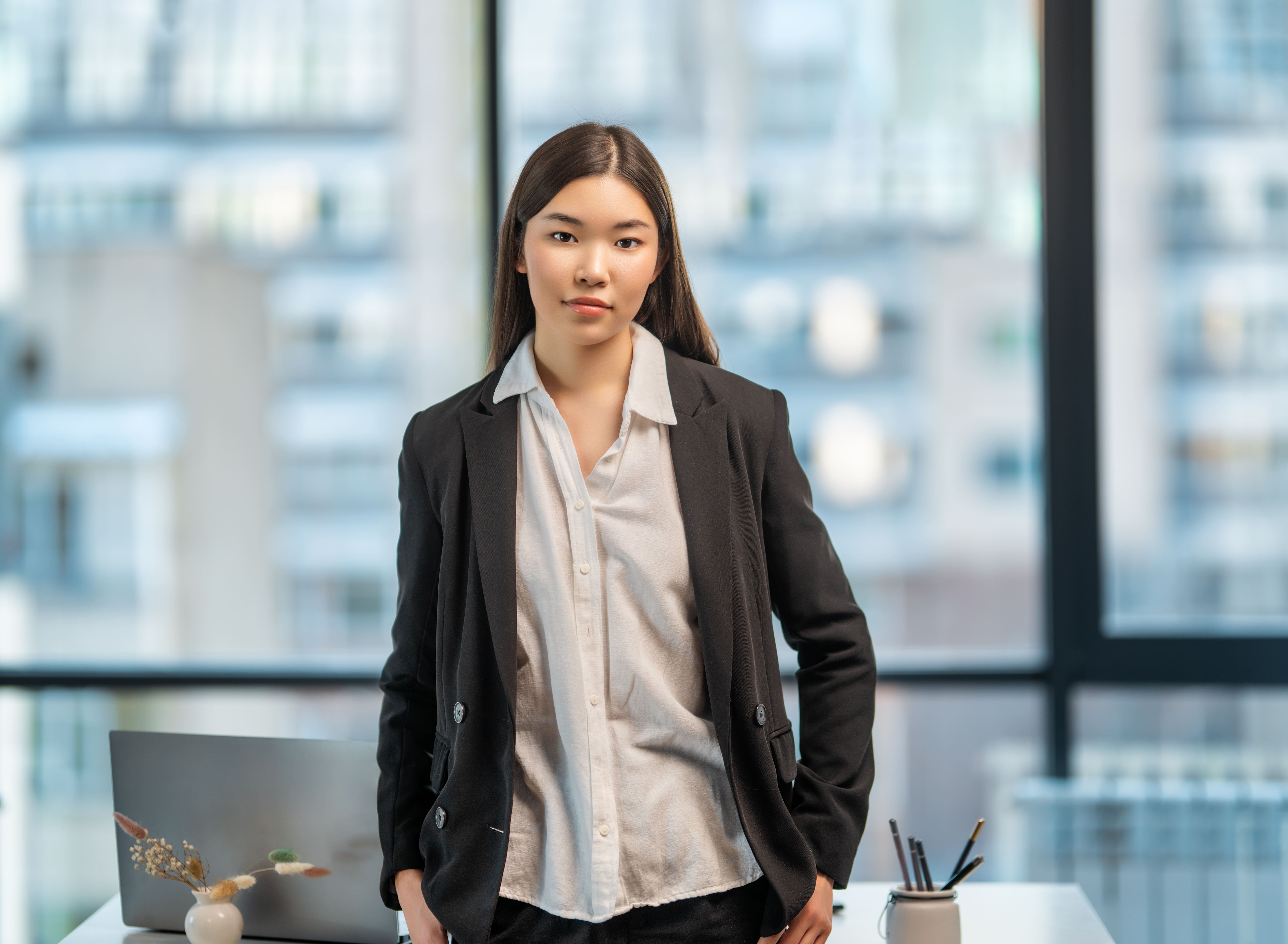 A woman in a black suit and white shirt standing in front of a window.