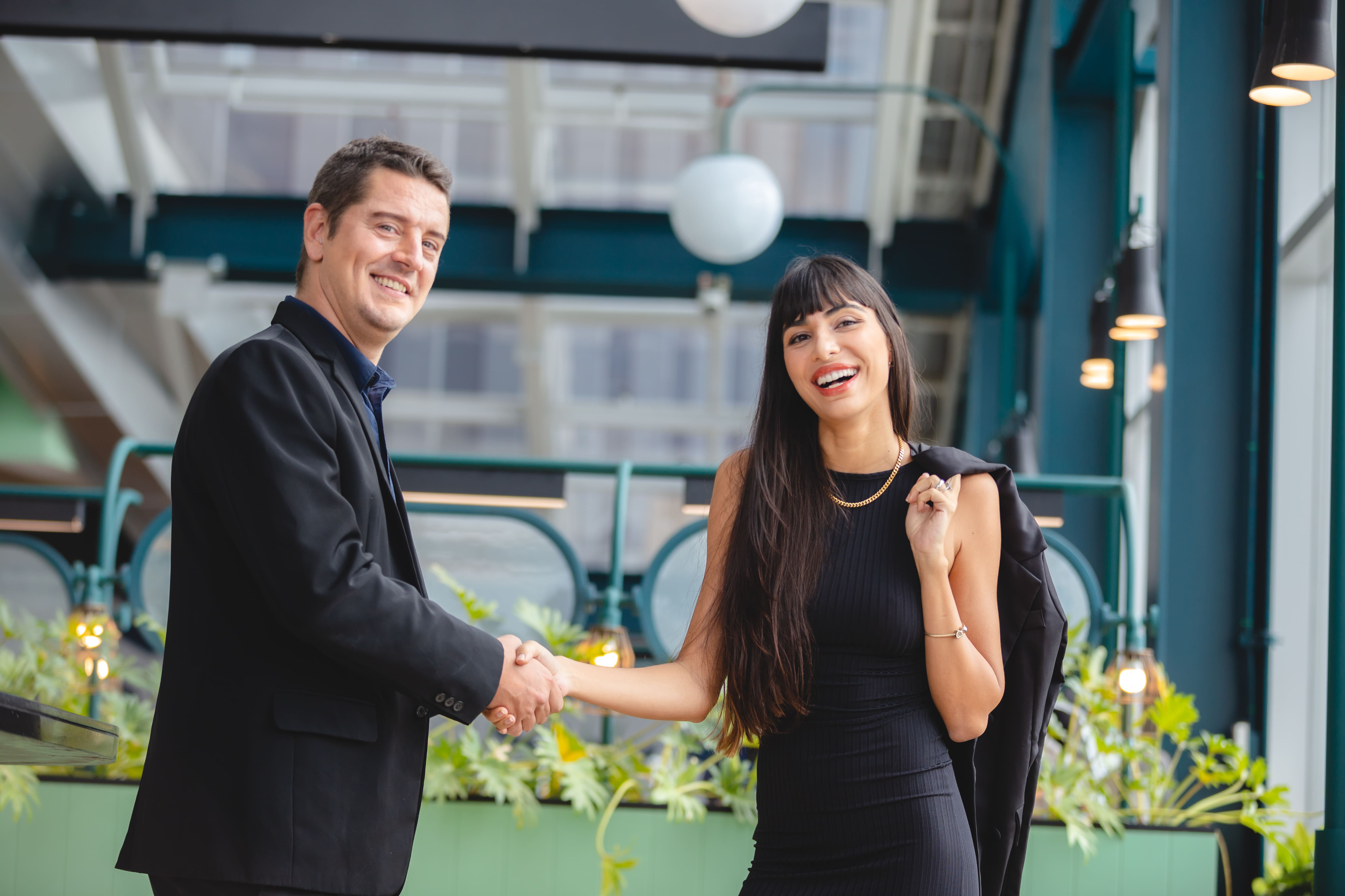 A man and a woman shake hands in a room with a green plant.