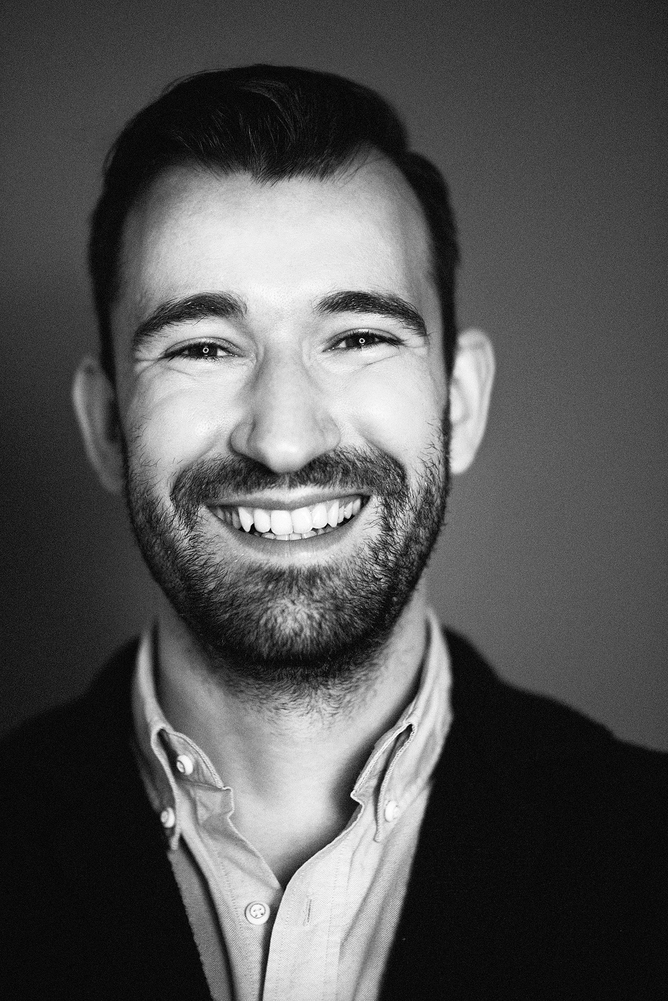 Black and white portrait of a cheerful man with a full beard, wearing a collared shirt and blazer, smiling confidently