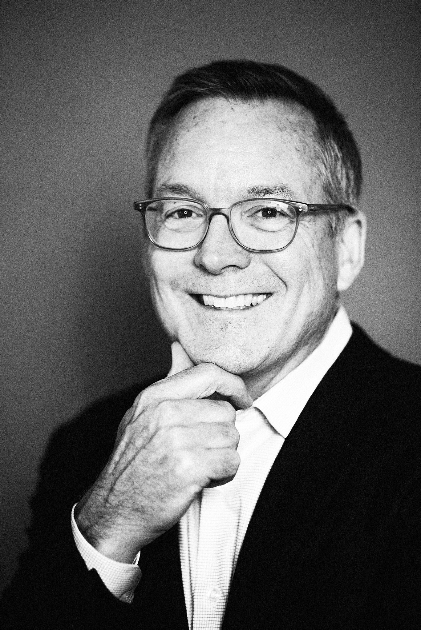 Black and white portrait of an older man wearing glasses, smiling confidently with hand resting on his chin