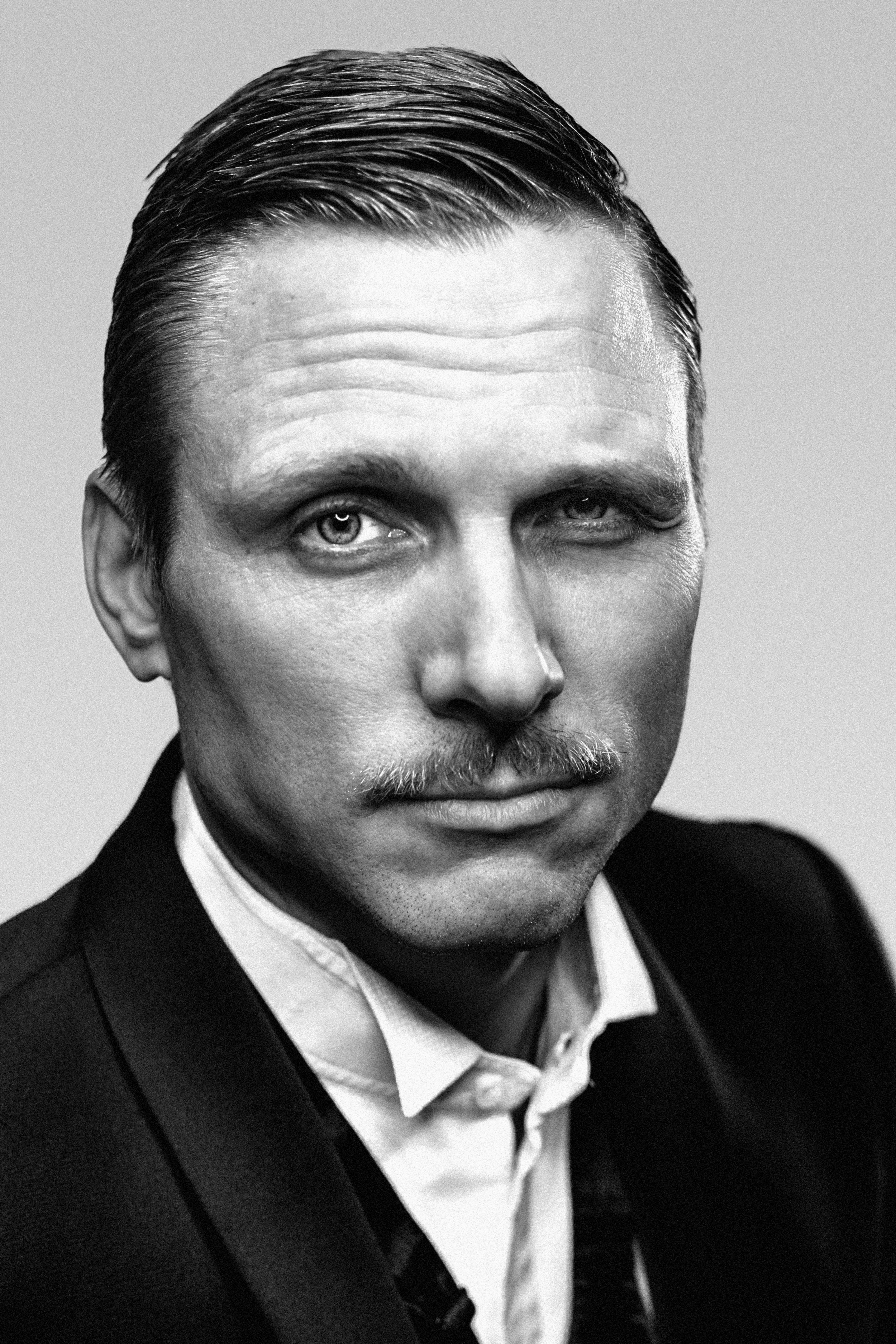 Black and white portrait of a man with a slicked-back hairstyle and mustache, wearing a suit and gazing intently at the camera