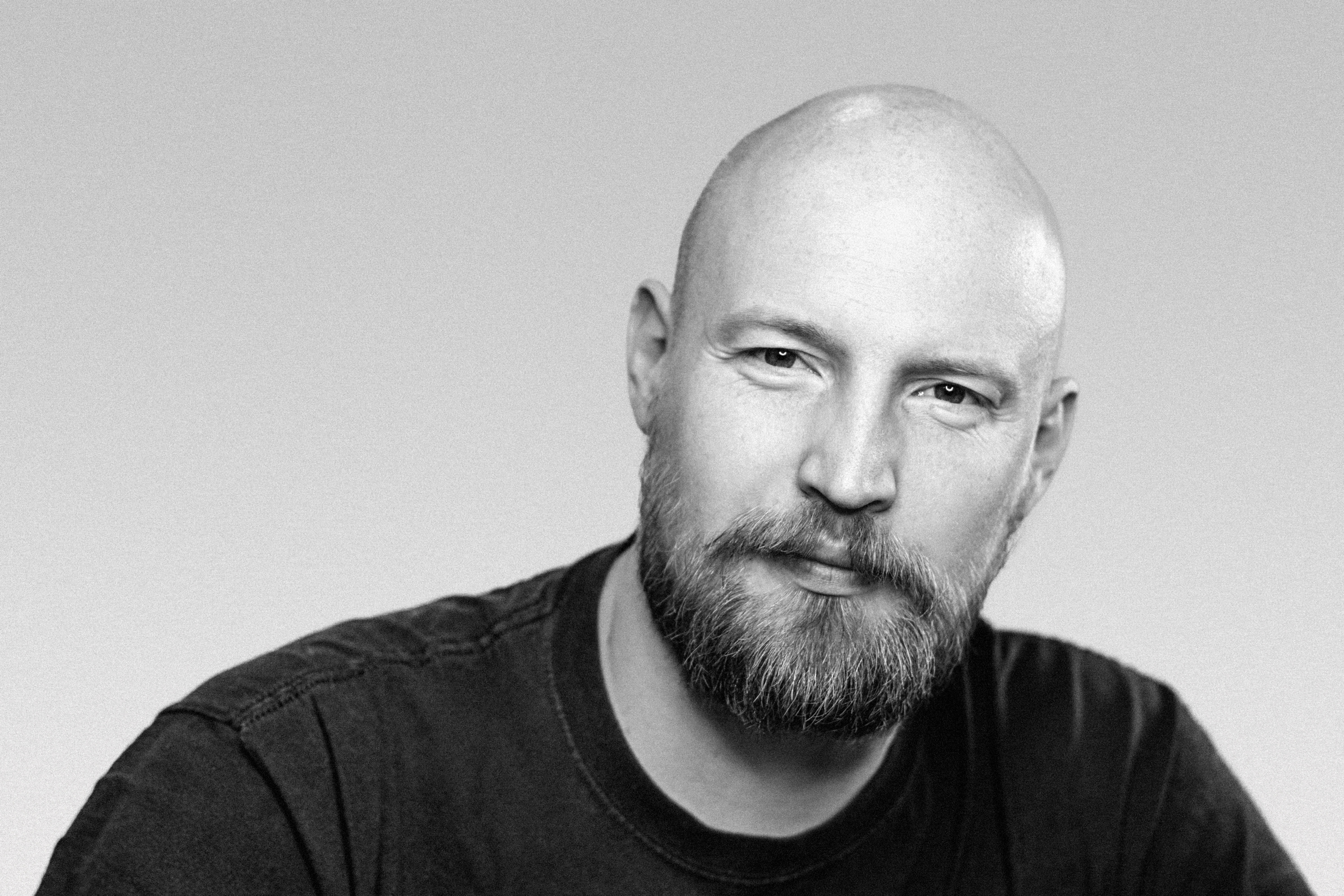 Black and white portrait of a bald man with a beard, wearing a dark shirt, looking directly at the camera