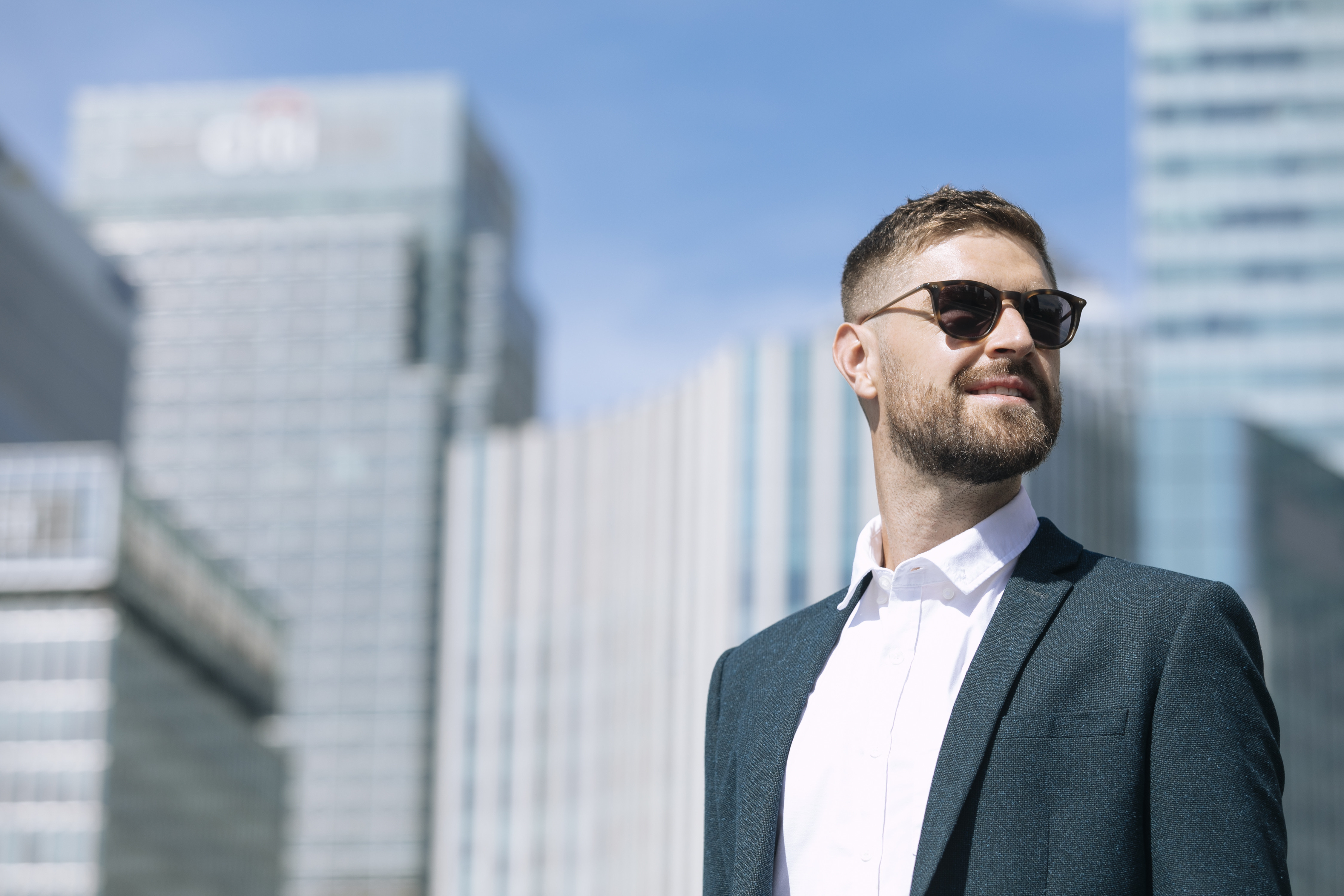 A man wearing sunglasses and a suit stands in front of a building.