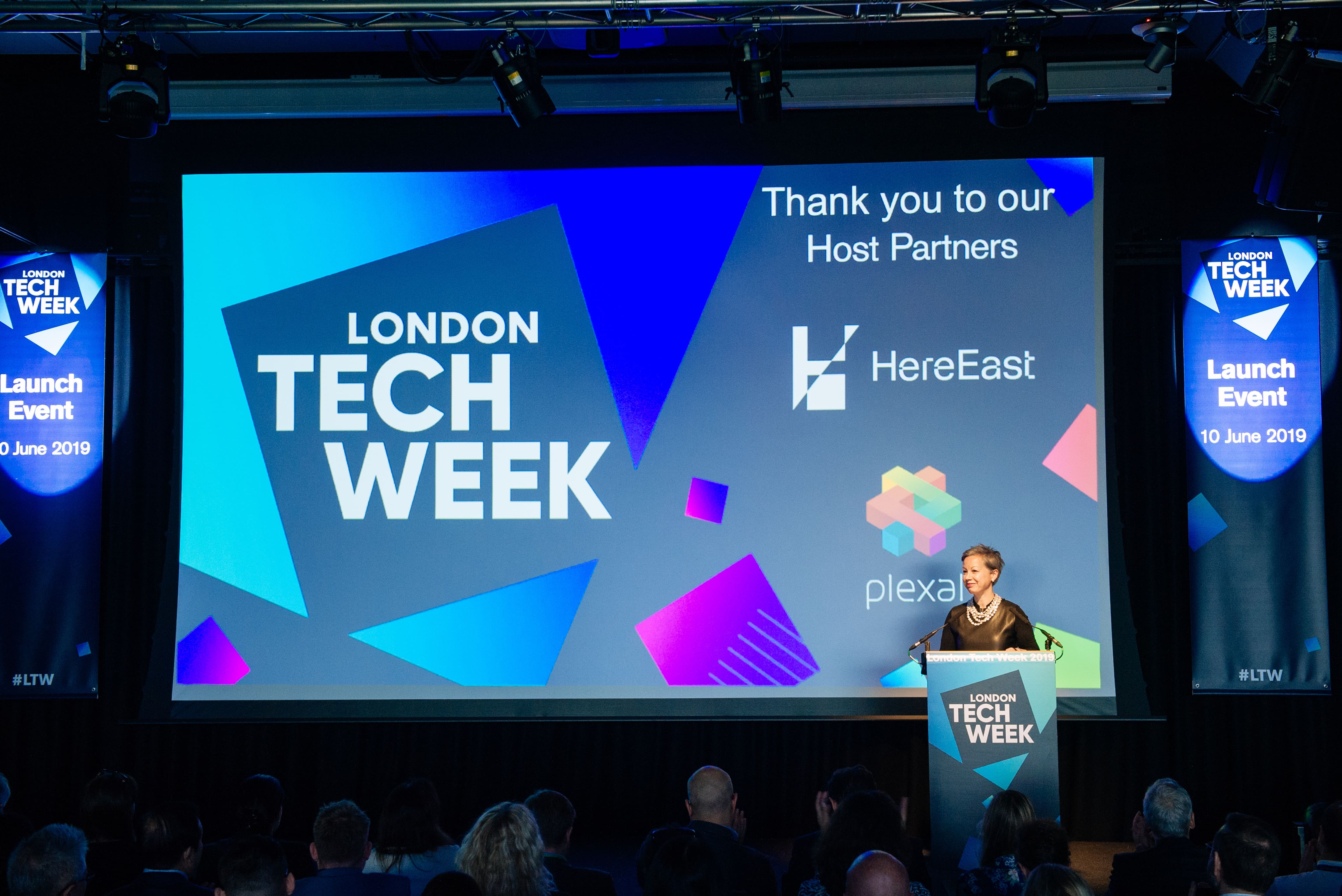 Speaker presenting at the London Tech Week 2019 launch event, with host partners HereEast and Plexal logos displayed on the screen