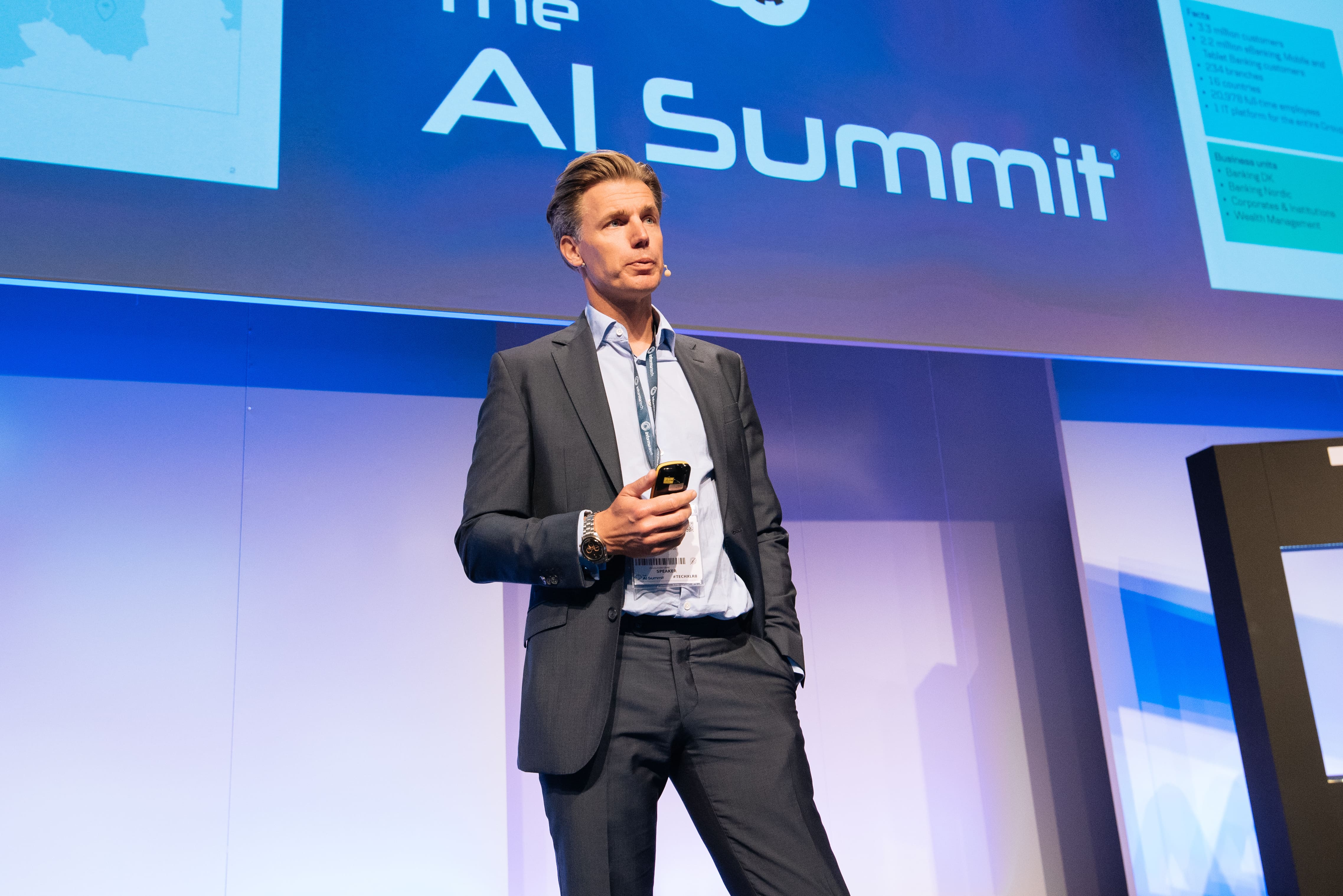 Speaker presenting at The AI Summit, standing on stage with a large digital screen displaying event branding