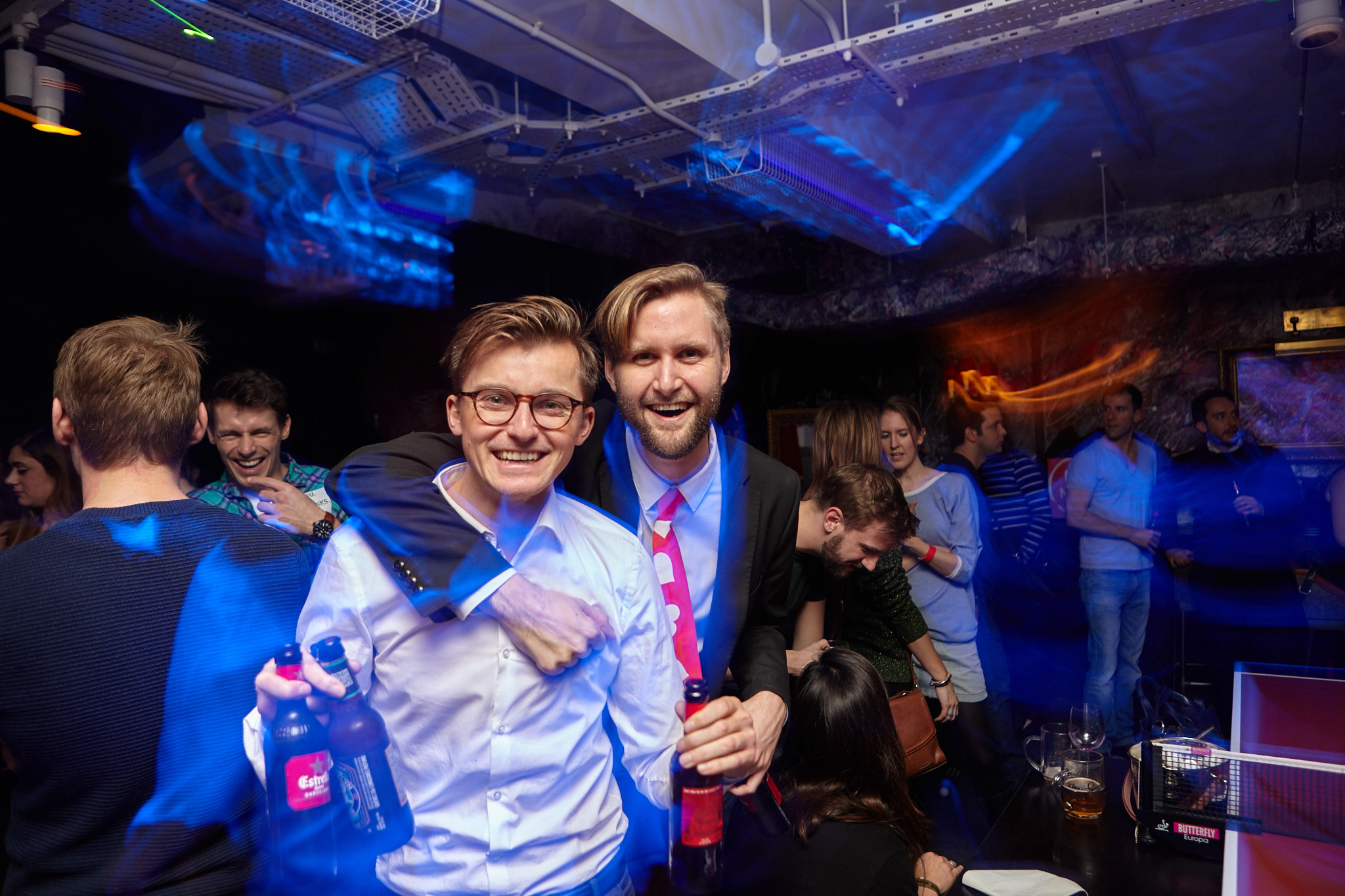 Two men in a bar with one holding a bottle of beer.