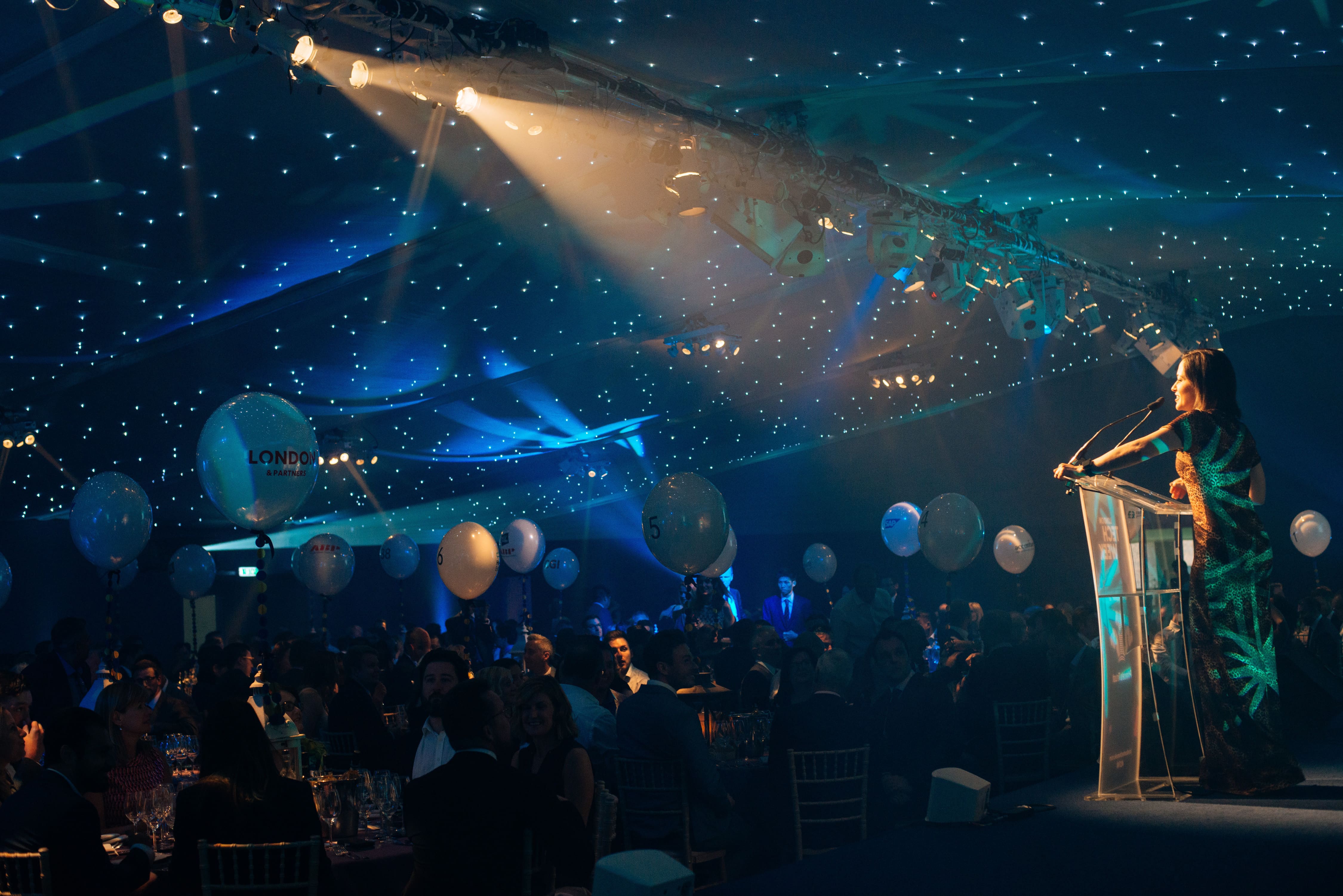 A large group of people are gathered in a room with a blue carpet and balloons.