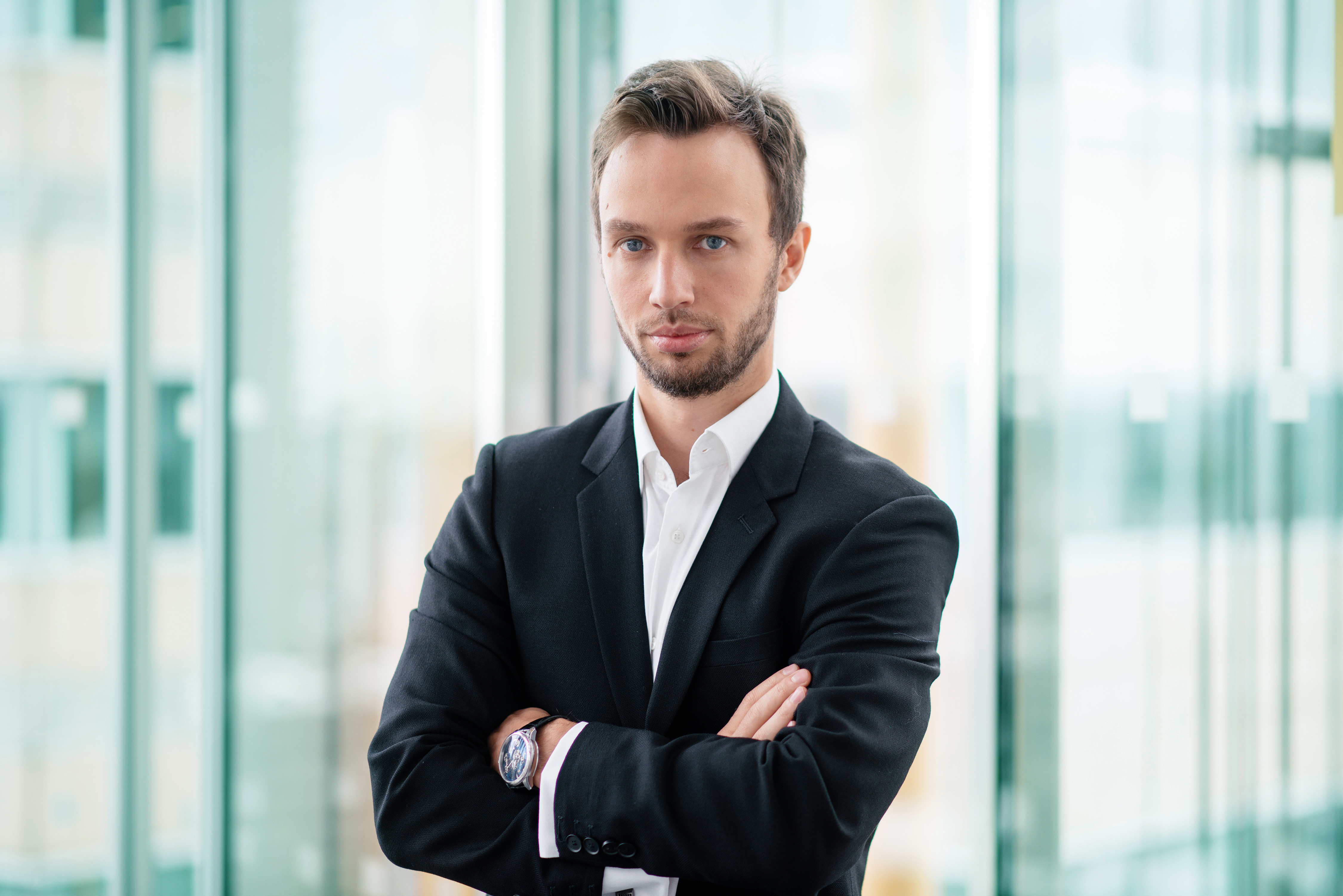 Professional headshot of a man in a black suit with arms crossed, standing in front of a glass-walled office space