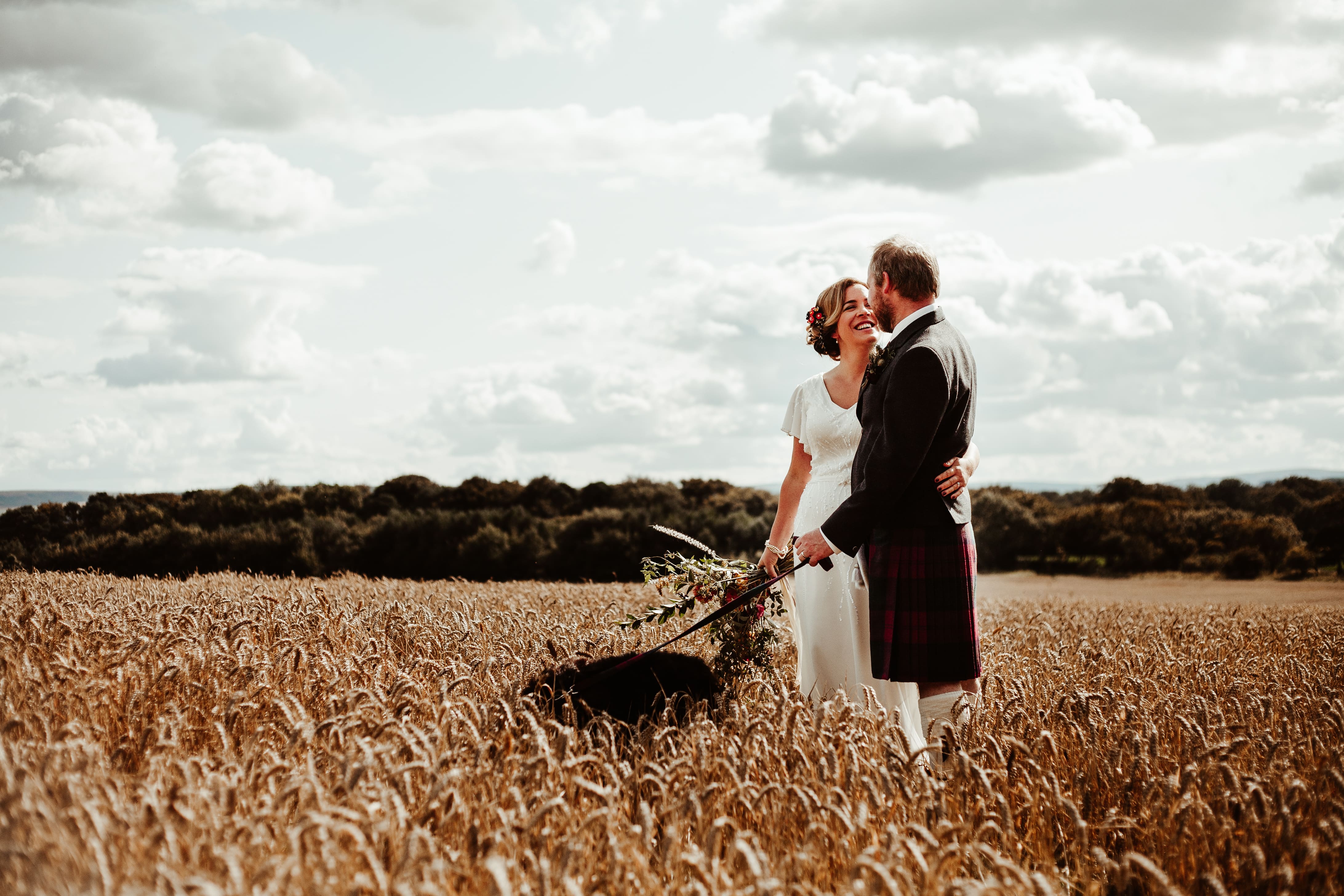 A bride and groom stand in a field with a black dog.