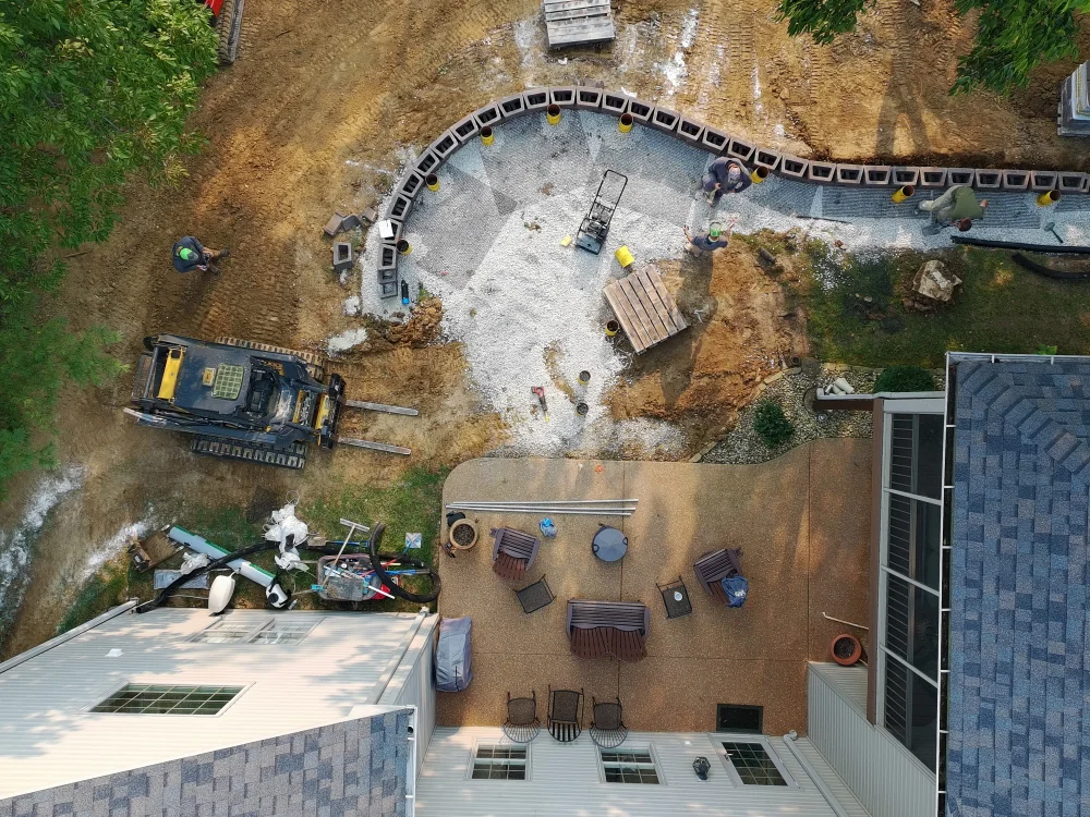 Aerial view of a backyard patio with construction work underway for a curved retaining wall surrounded by equipment and workers.